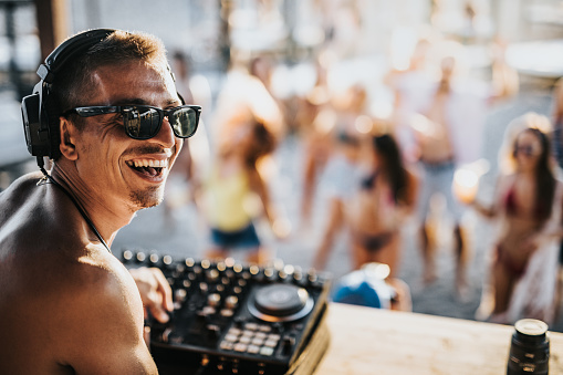 Young happy DJ playing music during a party on the beach and turning towards the camera.