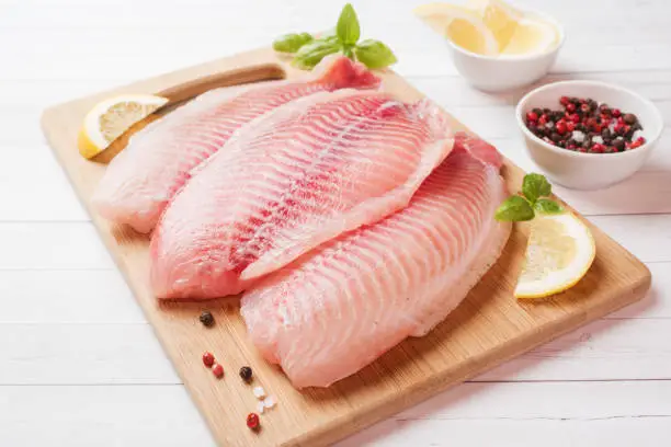 Raw fish fillet of tilapia on a cutting Board with lemon and spices. White table with copy space