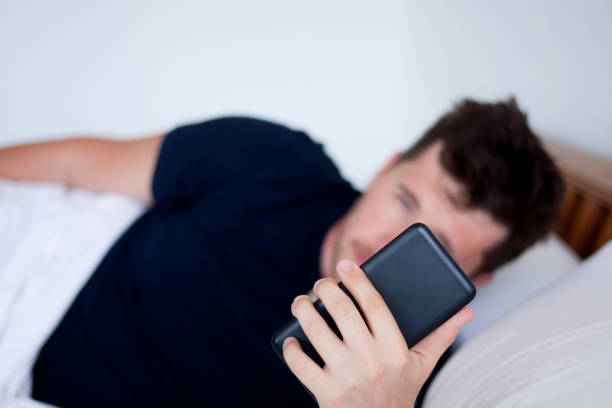 Sleepy caucasian young man lying in bed after waking up and using smartphone. Morning hangover. Early awakening with alarm clock on phone. stock photo