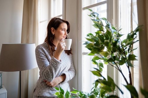 Mid adult woman having coffee in pajamas and looking through the window