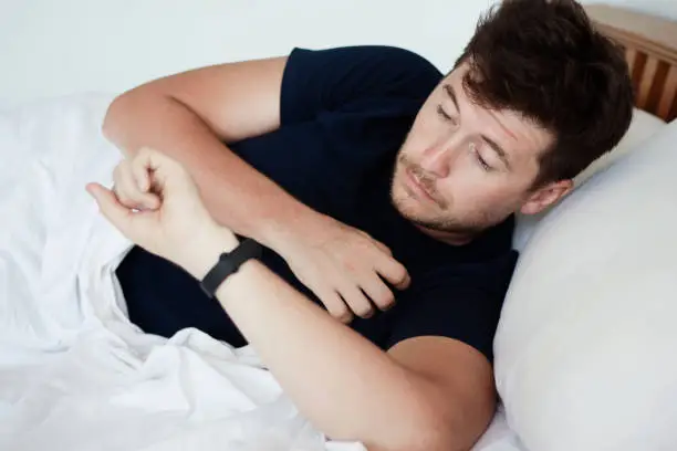 Sleepy young handsome caucasian man awake in early morning in his badroom and looking at alarm watches or fitness-tracker. Troubles with healthy sleep and tired after night sleep concept.