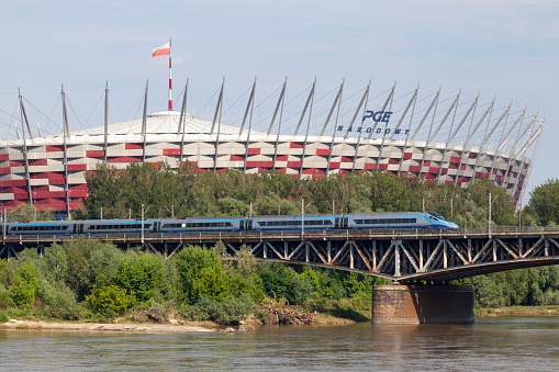 Warsaw, Poland - June 08 2019: A PKP Intercity ED250 Pendolino high-speed train crossing the Średnicowy bridge over the Vistula River with behind, the National Stadium.