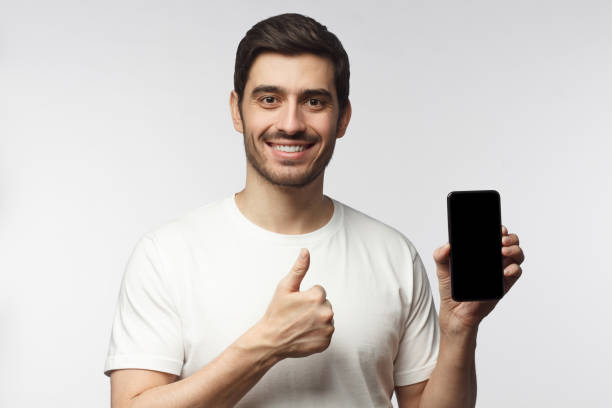 istock Attractive happy young man holding blank smartphone, smiling at camera, showing thubms up gesture, isolated on gray background 1155161596