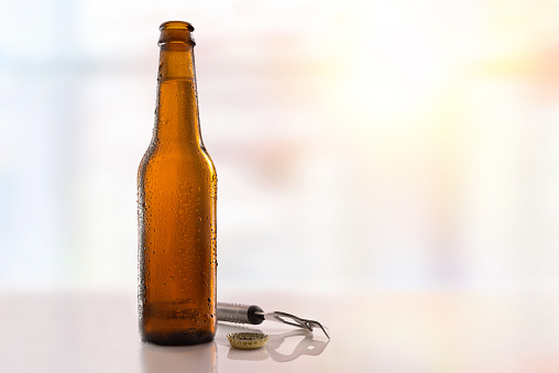 Beer bottle filled and open on glass table with bottle opener and light background. Horizontal composition. Front view.