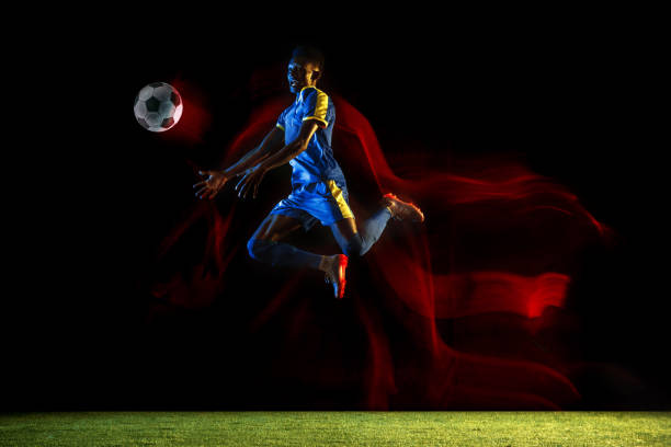 Young african-american male football or soccer player in sportwear and boots kicking ball for the goal in mixed light on dark background. Concept of healthy lifestyle, professional sport, hobby.