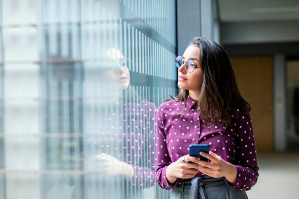 Female business professional taking a break from work Shot of a young female business professional with a cell phone standing by a glass wall in office and looking away soul searching stock pictures, royalty-free photos & images