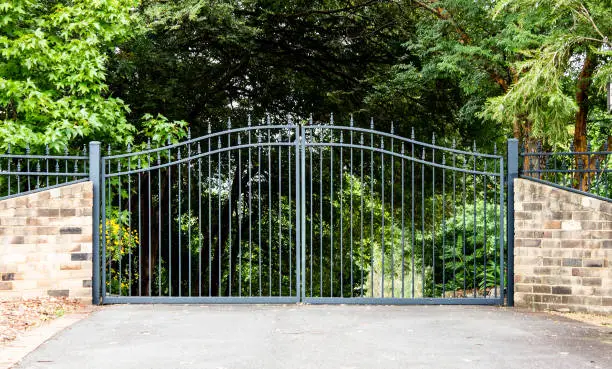 Photo of Metal driveway property entrance gates set in brick fence with garden trees  in background