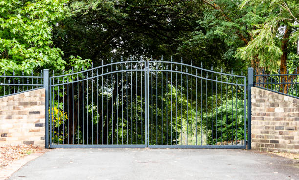 металлическая подъездная дорога собственности входные ворота, установленные в кирпичный забор с садовыми деревьями в фоновом режиме - ворота стоковые фото и изображения