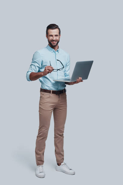 biznes to jego życie. pełna długość przystojnego młodego człowieka niosącego laptopa i patrzącego na kamerę stojąc na szarym tle - smart casual looking at camera full length indoors zdjęcia i obrazy z banku zdjęć