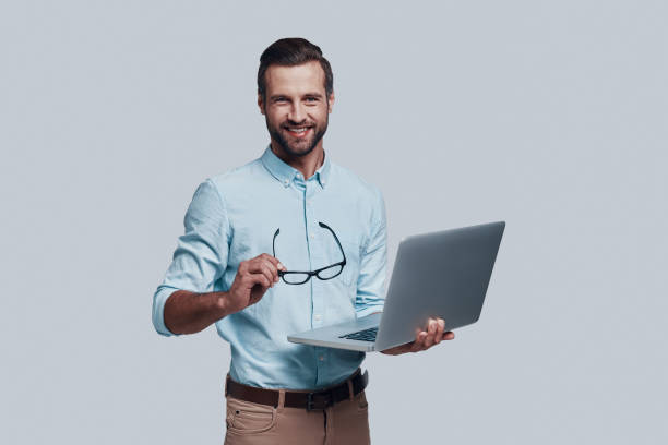 istock May I help you? Good looking young man carrying laptop and looking at camera while standing against grey background 1155173870