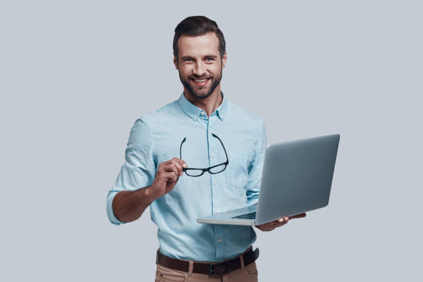 istock Always ready to help. Good looking young man carrying laptop and looking at camera while standing against grey background 1155173933