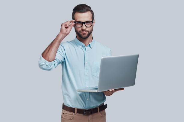 istock Modern technologies. Good looking young man carrying laptop and looking at camera while standing against grey background 1155173988