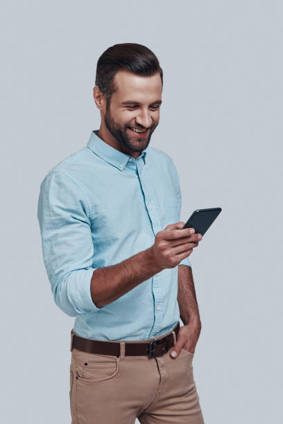 istock Receiving good feedbacks. Handsome young man using smart phone and smiling while standing against grey background 1155175089