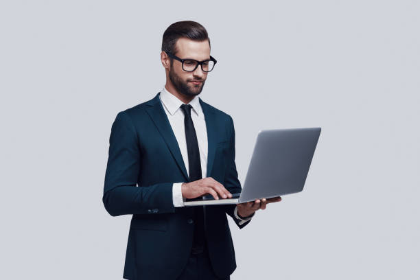 istock Paying attention to every detail. Handsome young man using laptop and smiling while standing against grey background 1155176249