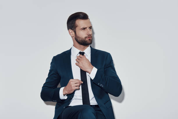 istock Paying attention to every detail. Handsome young man adjusting his necktie while sitting against grey background 1155178970