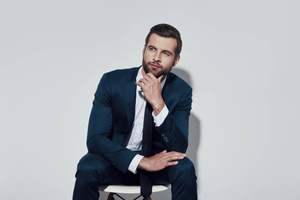 istock Solution is near. Handsome young man looking away and keeping hand on chin while sitting against grey background 1155179243