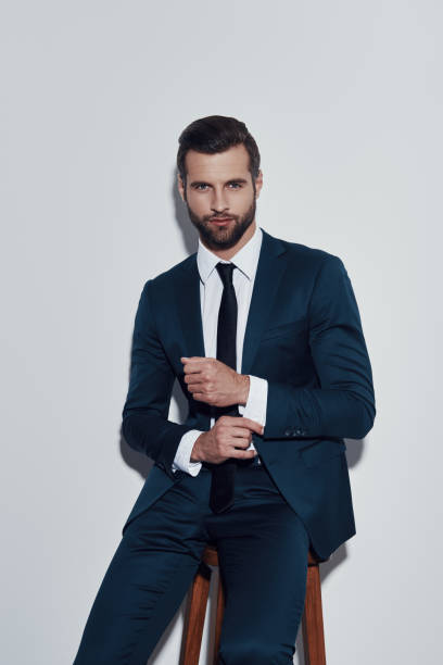 istock Irresistible man. Handsome young man adjusting his sleeve and looking at camera while sitting against grey background 1155179624