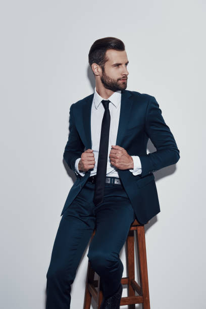 istock Charming man. Handsome young man adjusting his jacket and looking away while sitting against grey background 1155179656
