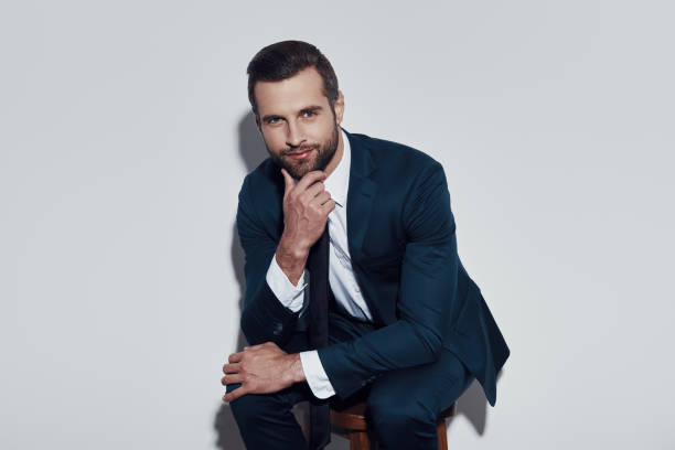 istock Taking time to think. Handsome young man looking at camera with smile and keeping hand on chin while sitting against grey background 1155180177