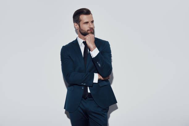istock Lost in thoughts. Charming young man keeping hand on chin and smiling while standing against grey background 1155180285