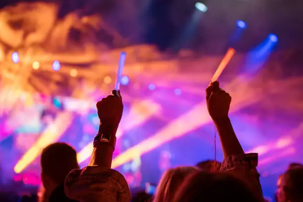 Photo of Young teenager girl fan raised up two hands with colored glowing sticks supporting favorite popular band on the night concert. Colorful crowdy background entertainment with light and laser show.