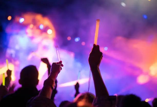 Photo of Young teenager girl fan raised up two hands with colored glowing sticks supporting favorite popular band on the night concert. Colorful crowdy background entertainment with light and laser show.