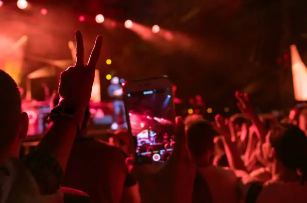 Photo of Teenager girl fan raised hand with V sign Gesture and shooting vertical video with smartphone popular band on the night concert. Colorful crowdy background entertainment with light and laser show.
