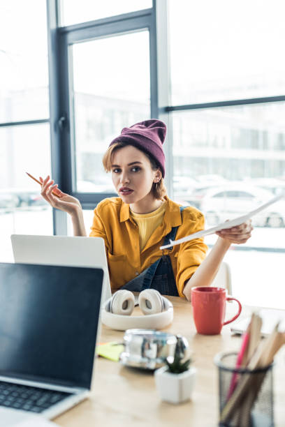 istock confused young female it specialist holding document and pen while sitting at computer desk in loft office 1155183801
