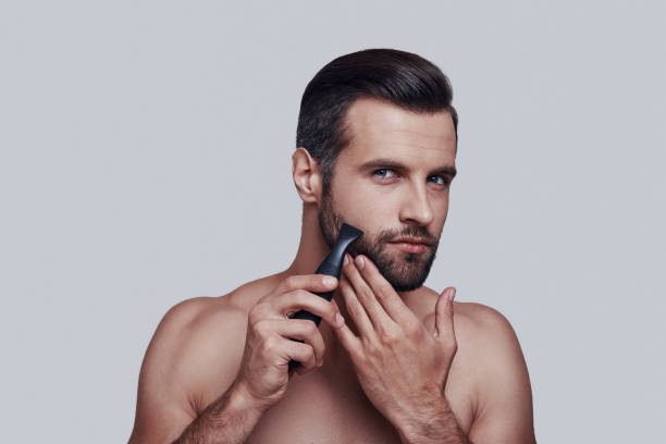 istock Taking care of his beard. Handsome young man shaving and looking at camera while standing against grey background 1155187767