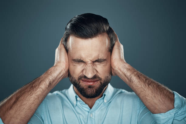 problemas. joven frustrado cubriendo las orejas con las manos mientras se está parado contra el fondo gris - hand to ear fotografías e imágenes de stock