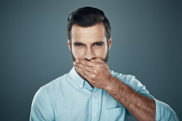 istock Silence. Handsome young man looking at camera and covering mouth with hand while standing against grey background 1155191385