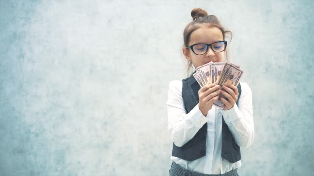 The small business lady stands on a gray background with dollars in her hands. During this, she waves a bundle of money, smiles at the camera. Smiles.