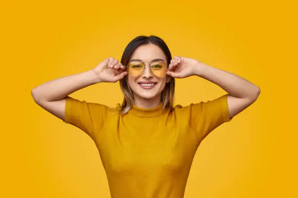 Young lady in trendy yellow T-shirt adjusting sunglasses and smiling for camera against vivid background