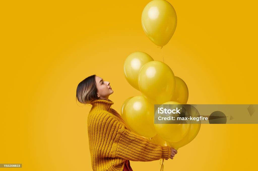Young woman releasing balloons Side view of young female in knitted sweater looking up and releasing bunch of bright balloons during birthday party against yellow background Balloon Stock Photo