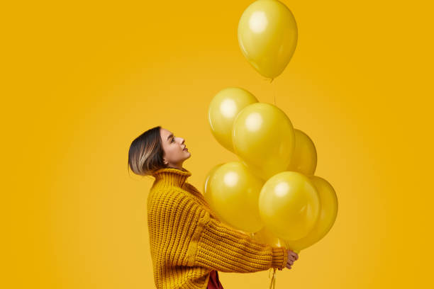 Young woman releasing balloons Side view of young female in knitted sweater looking up and releasing bunch of bright balloons during birthday party against yellow background releasing stock pictures, royalty-free photos & images
