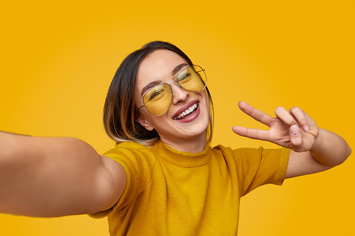 Young female in stylish sunglasses cheerfully smiling and showing V sign while taking selfie against yellow background