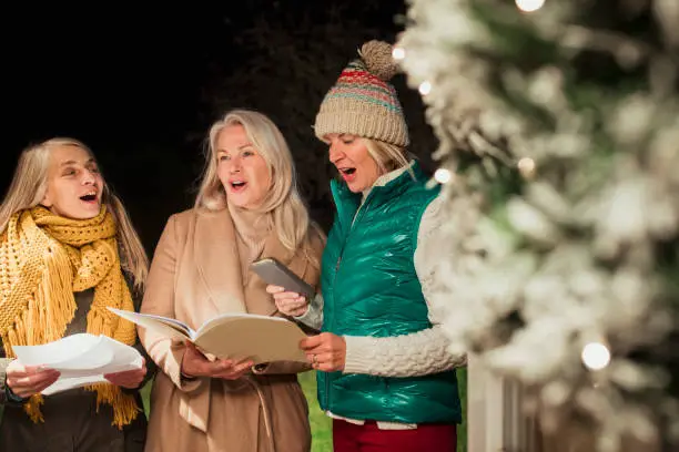 Group of three women dressed warmly standing at a front door holding books as they Christmas carol.