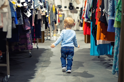 Cute caucasian blond toddler boy walking alone at clothes retail store between rack with hangers. Baby discovers adult shopping world. Baby get lost at big hypermarket shopping mall.