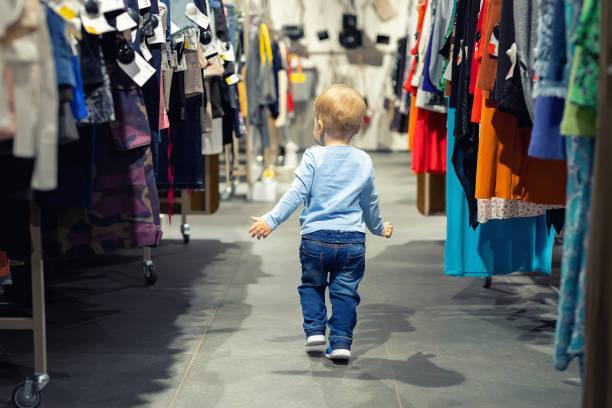 Cute caucasian blond toddler boy walking alone at clothes retail store between rack with hangers. Baby discovers adult shopping world. Baby get lost at big hypermarket shopping mall stock photo