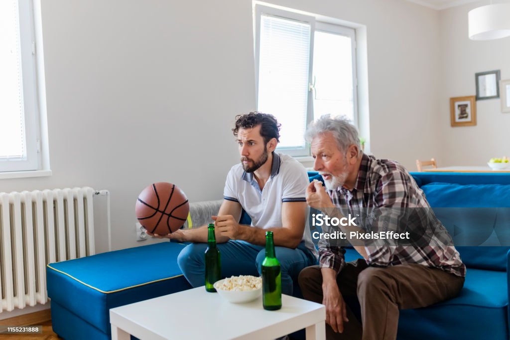 Sport brings family together Senior Father and Adult Son Basketball Fans Watching Basketball Game on Tv in Their Livingroom. Elderly Man Watching Championship With Handsome Son With Basketball in Their Hands. Family, Sports and Entertainment Concept. 25-29 Years Stock Photo