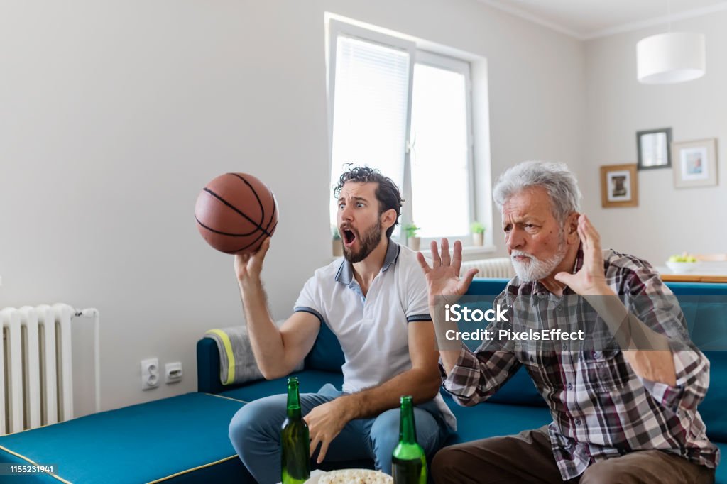 Epic, exciting game! Senior Father and Adult Son Basketball Fans Watching Basketball Game on Tv in Their Livingroom. Elderly Man Watching Championship With Handsome Son With Basketball in Their Hands. Family, Sports and Entertainment Concept. Basketball - Sport Stock Photo