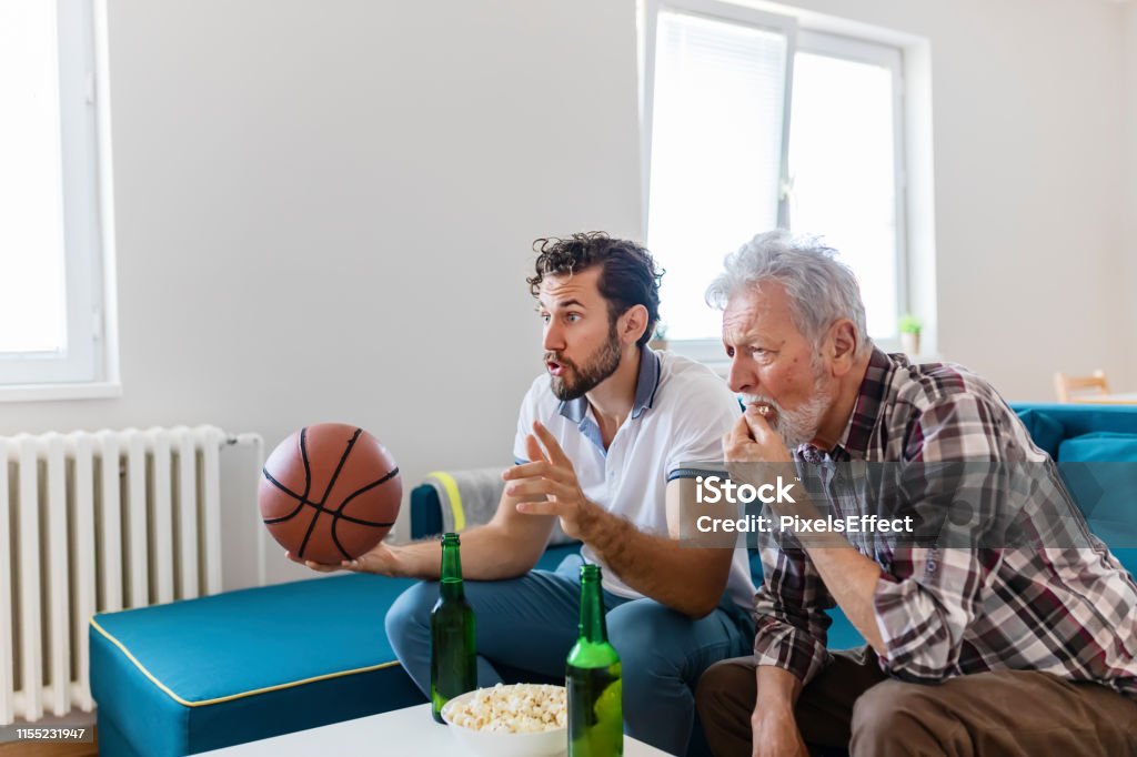 Sport brings family together Senior Father and Adult Son Basketball Fans Watching Basketball Game on Tv in Their Livingroom. Elderly Man Watching Championship With Handsome Son With Basketball in Their Hands. Family, Sports and Entertainment Concept. 25-29 Years Stock Photo