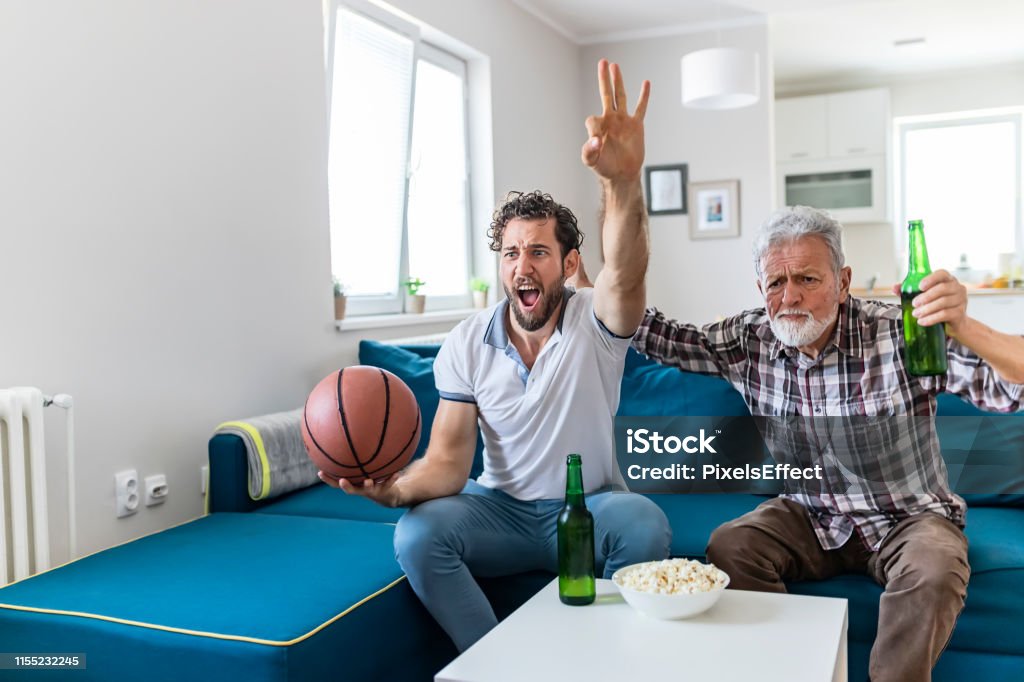 Wow, what a game... Senior Father and Adult Son Basketball Fans Watching Basketball Game on Tv in Their Livingroom. Elderly Man Watching Championship With Handsome Son With Basketball in Their Hands. Family, Sports and Entertainment Concept. 25-29 Years Stock Photo