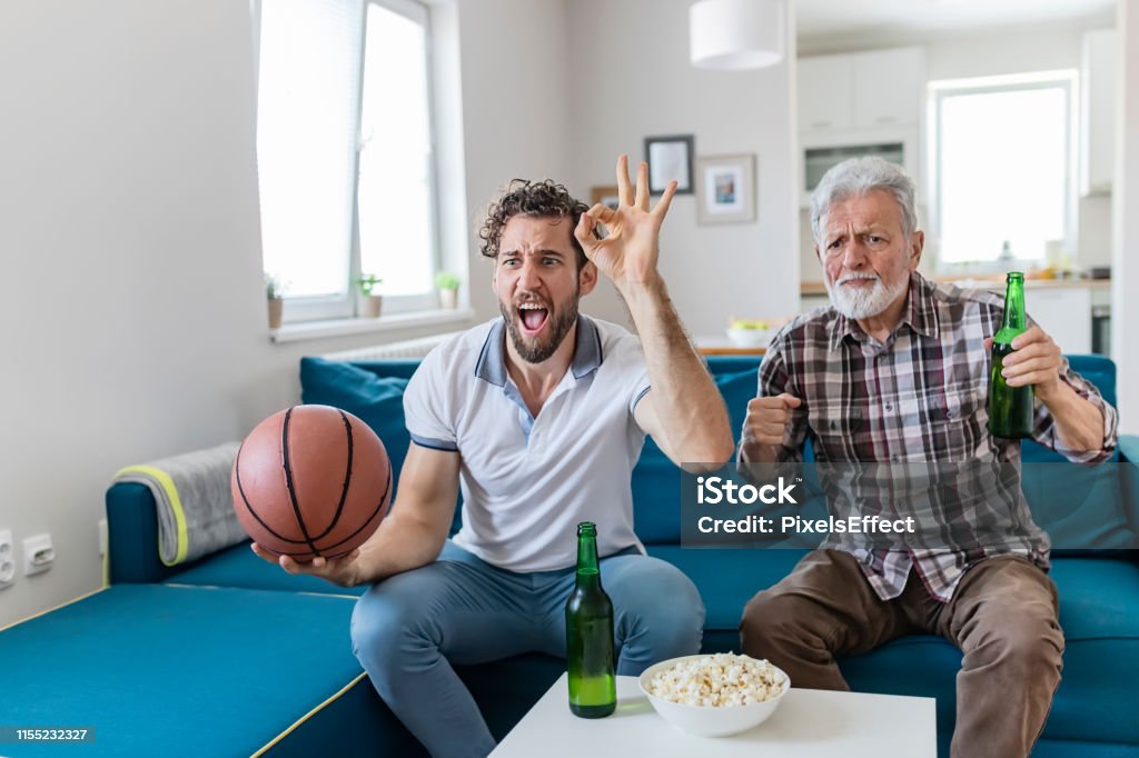 Wow, what a game... Senior Father and Adult Son Basketball Fans Watching Basketball Game on Tv in Their Livingroom. Elderly Man Watching Championship With Handsome Son With Basketball in Their Hands. Family, Sports and Entertainment Concept. 25-29 Years Stock Photo