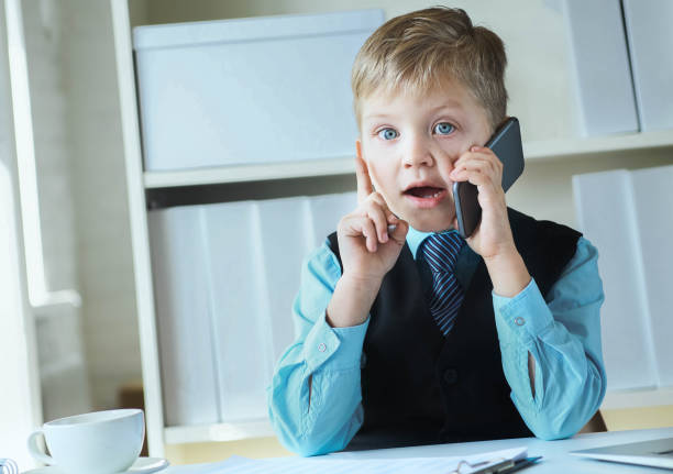 istock Little confident executive businessman boss boy making notes and talks on the phone sitting at the desk with laptop in office. 1155236554