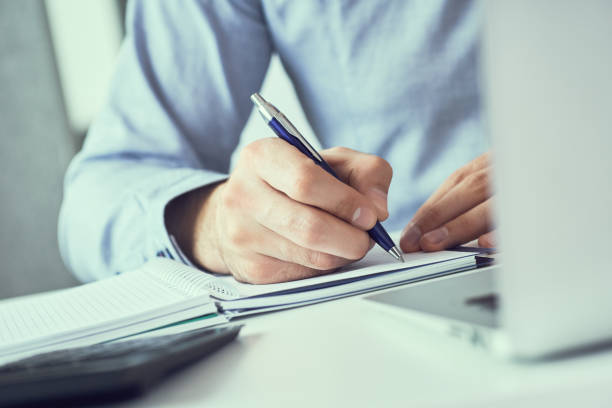 istock Businessman hand writing note on a notebook. Business man working at office desk. Close up of empty notebook on a blackboard with office supplies. 1155236571