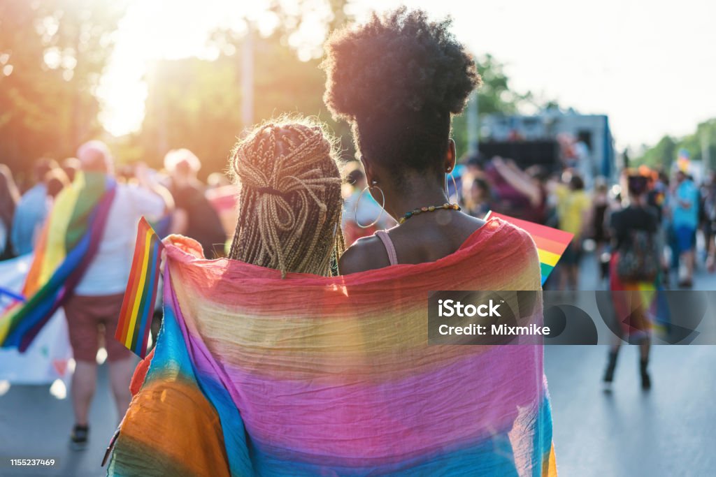 Young female couple hugging with rainbow scarf at the pride event Rear view image of young couple walking with the pride event, hugging and waving pride flags LGBTQIA Pride Event Stock Photo