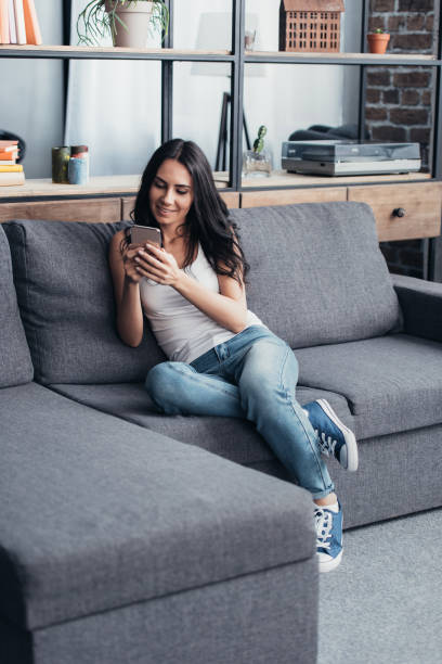 istock Smiling brunette woman with smartphone sitting on sofa 1155242500