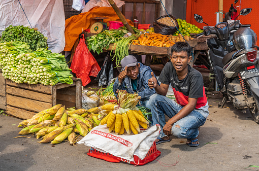 Makassar, Sulawesi, Indonesia - February 28, 2019: Terong Street Market. Two guys sell corn on the cob on side of vegetable booth. They flash positive hand signals.