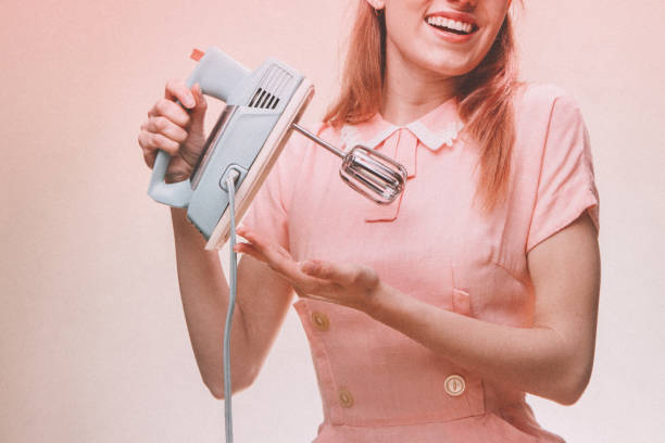 Retro Stereotypical Nineteen Fifties Portrait of Housewife A 1950's styled young woman in a vintage pink dress and cats eye glasses.  She hold up a vintage electric mixer, performing her duties as a good 50's wife.  Pink background. electric mixer photos stock pictures, royalty-free photos & images